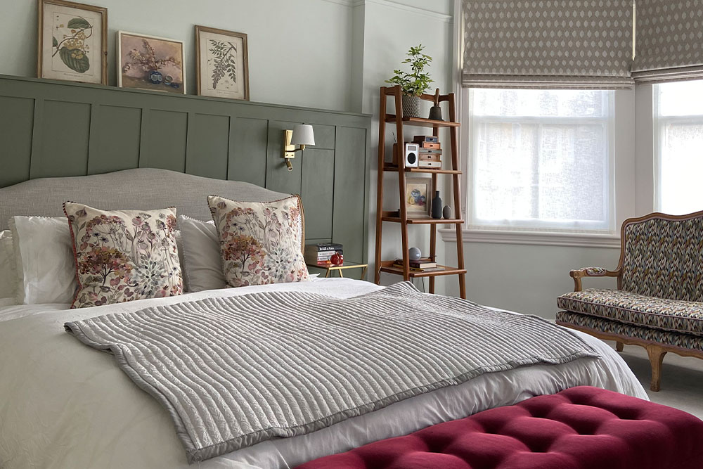 A cosy bedroom with a green panelled wall, a light grey bedspread, floral pillows, wooden shelf, and patterned furniture, creating a warm, inviting atmosphere.