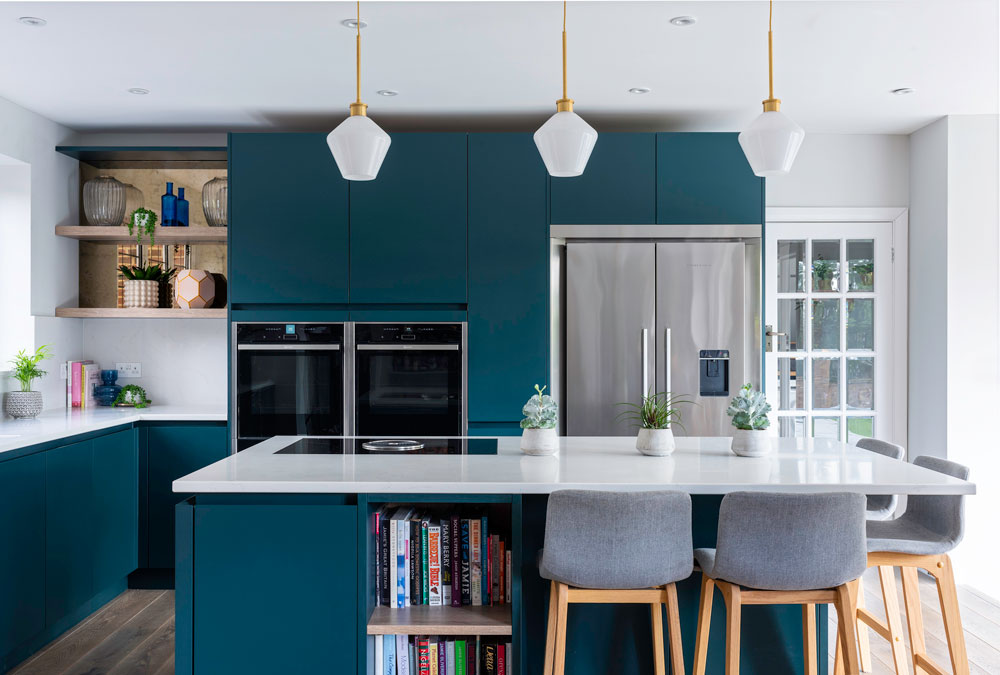Modern kitchen with teal cabinets, white countertops, and stainless steel fridge. Three pendant lights hang above the island with bar stools and plants.