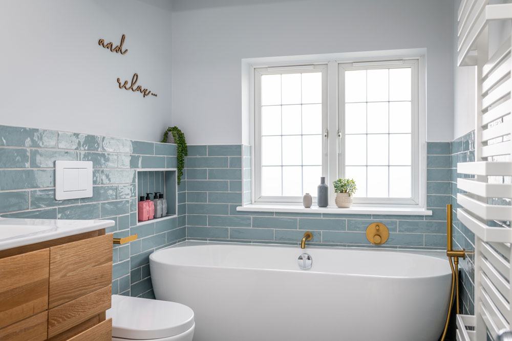 Modern bathroom with a white tub, gold fixtures, and light blue tiles. A wooden cabinet, window, and plants add a serene, relaxing feel.