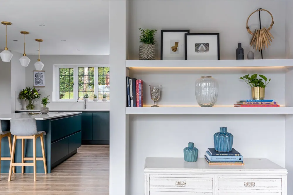 Spacious modern kitchen with teal cabinets, wooden stools, and pendant lights. Adjacent shelves display books, plants, and decorations, creating a cosy ambiance.