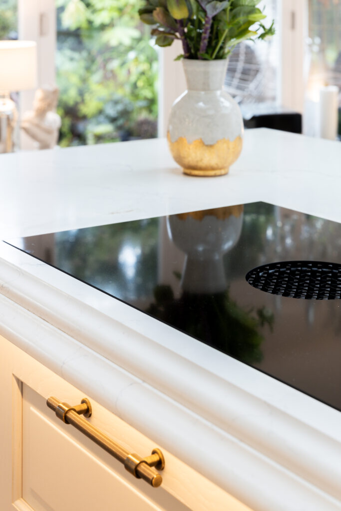 Modern kitchen with a sleek black induction cooktop on a white countertop. A vase with flowers adds a touch of nature, while brass handles enhance elegance.