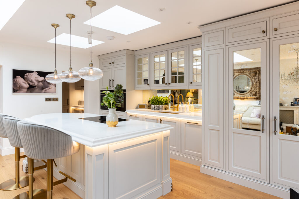 Bespoke Kitchens by Georgie Hall. Luxurious white kitchen with an island, pendant lights, and bar stools. Cabinets and mirrored doors reflect soft lighting, creating a cosy atmosphere.