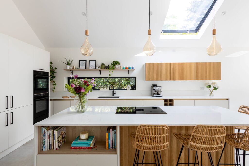 Bespoke Kitchens by Georgie Hall, modern kitchen with skylight, white cabinets, wood accents, and hanging bulbs. An island holds books and flowers, creating a bright, inviting space.