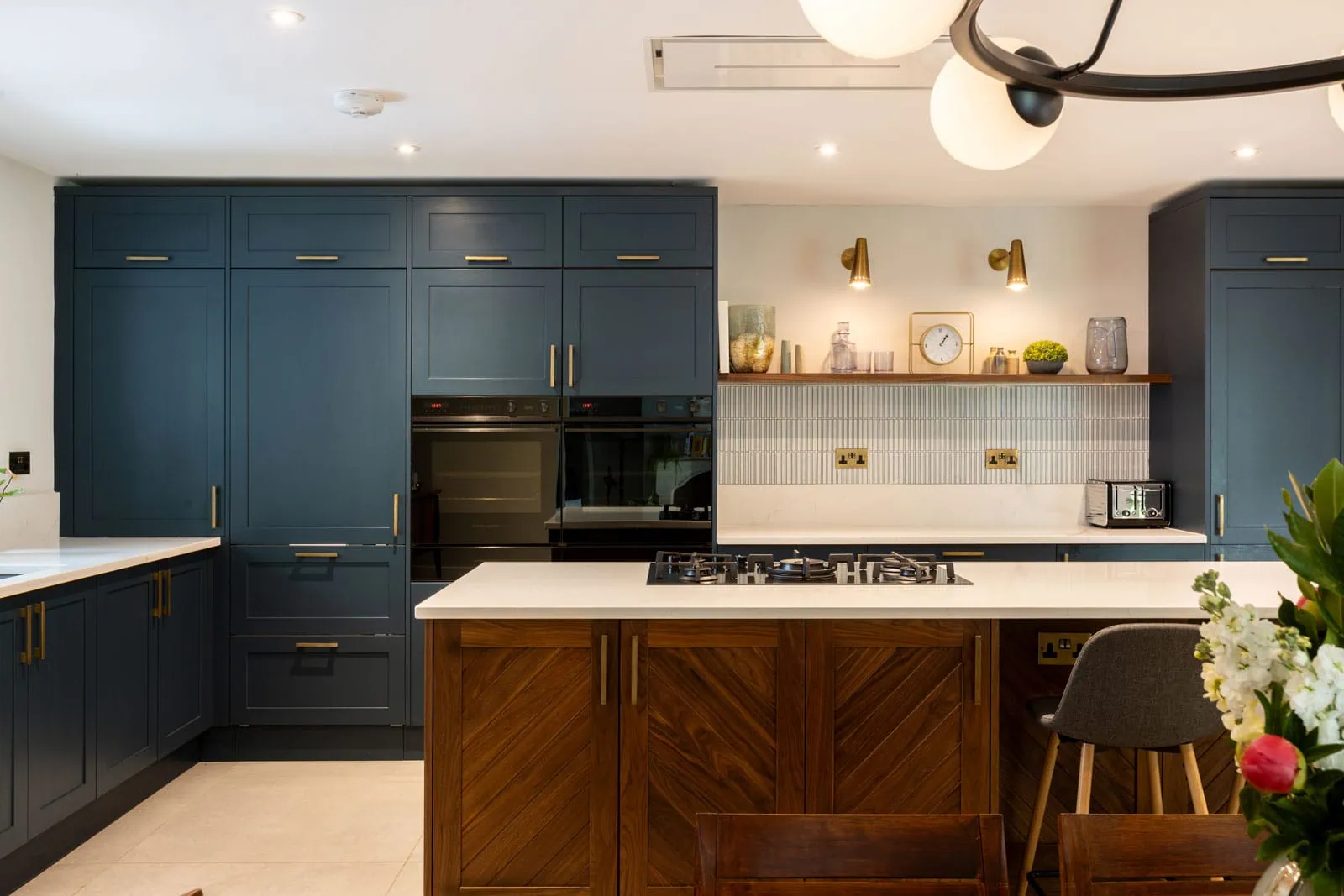 Modern kitchen with navy cabinets, wood-accented island, white countertops, and warm lighting. Shelves hold decorations and a clock, creating a cosy vibe.