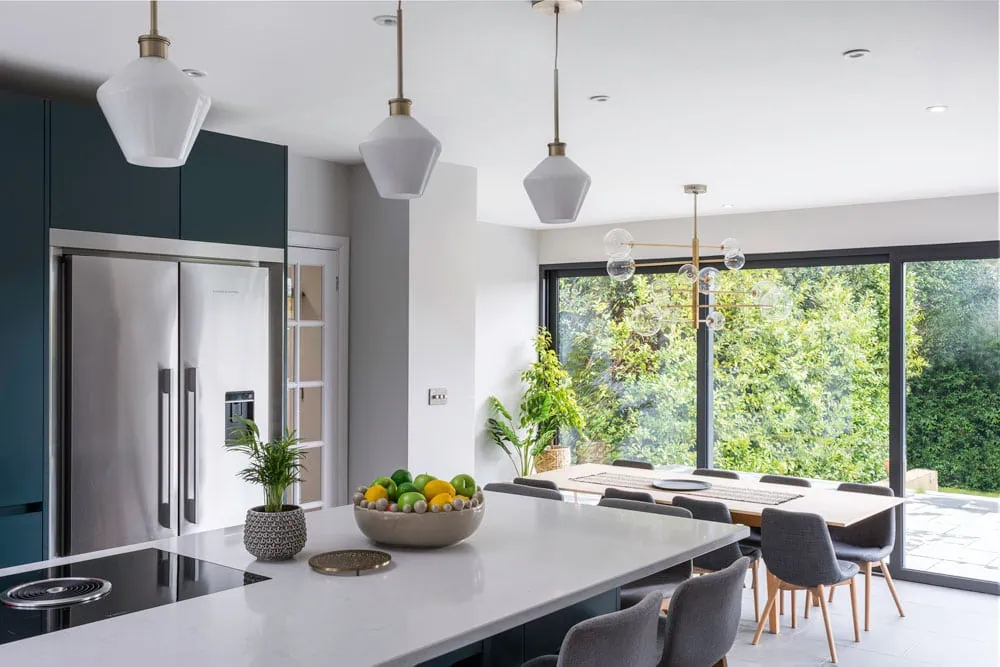 Modern kitchen with a sleek white island, pendant lights, and green cabinets. Large windows illuminate the dining area with garden views.
