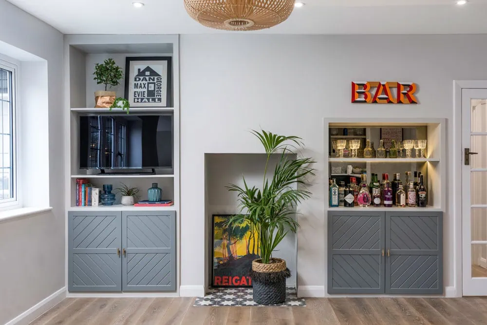 Contemporary living room with built-in shelves, grey cabinets, and a potted plant. A sign reads "BAR," and various bottles are displayed, creating a stylish, cosy vibe.