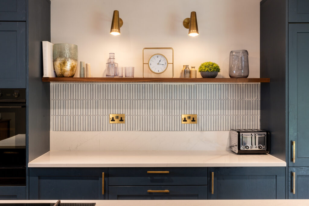 Bespoke Kitchens by Georgie Hall, modern kitchen with blue cabinets, marble countertop, and a finger tile backsplash. Warm lighting from brass sconces highlights a wooden shelf.
