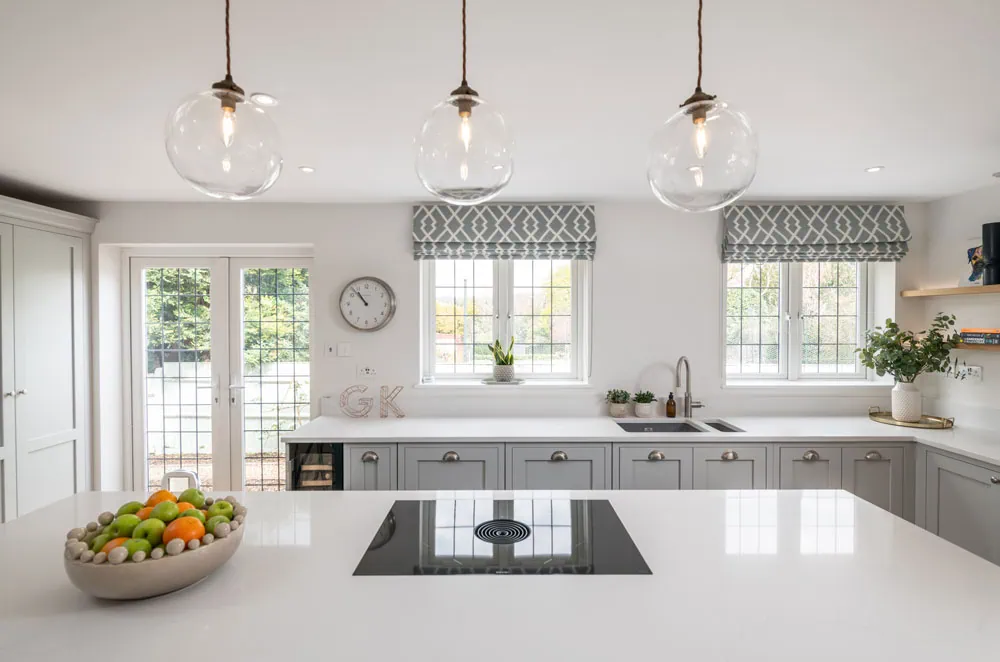 Bright kitchen with a modern design featuring a central island with a stovetop and fruit bowl. Large windows, hanging lights, and a cosy ambiance.