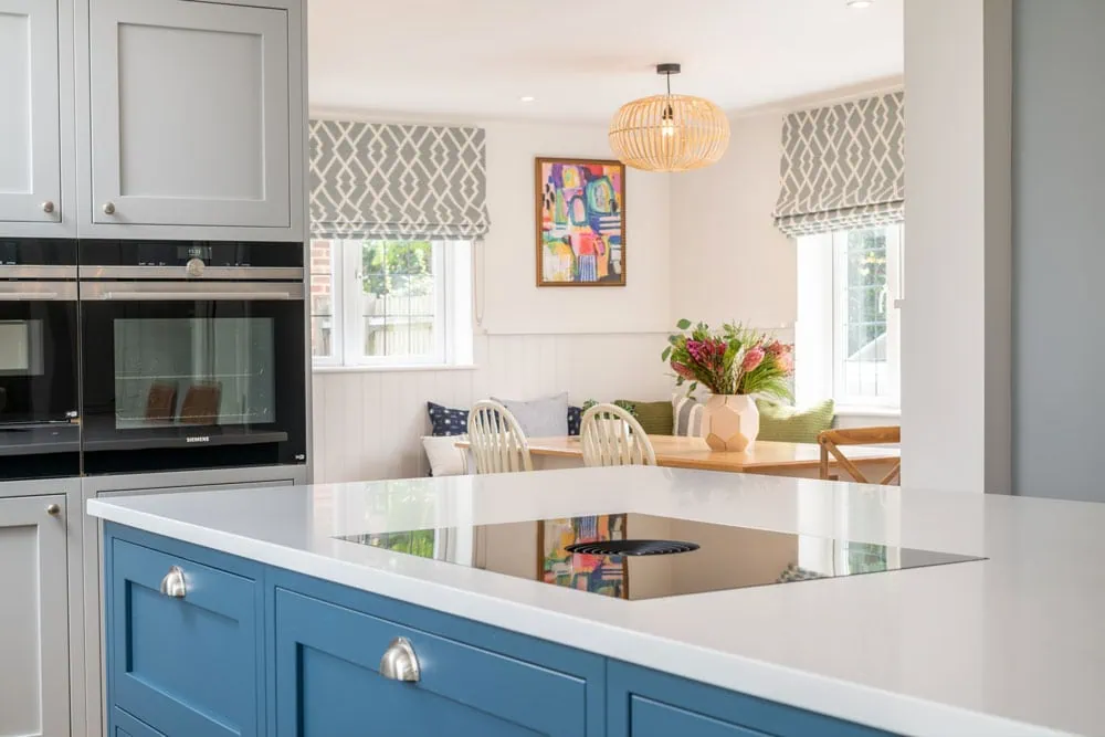 Modern kitchen with a sleek blue island and stovetop, adjacent to a cosy dining area. Warm, inviting tones and floral centrepiece enhance the space.
