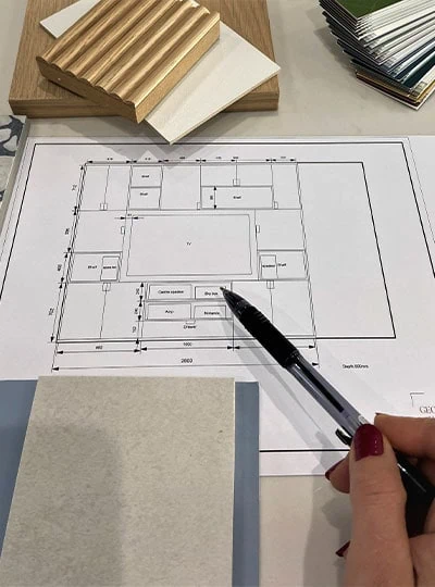 A person's hand points at a floor plan with a pen. Surrounding the plan are wood and paint samples, showing interior design planning.