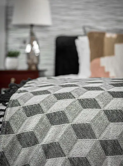 A soft, geometric-patterned blanket in grey tones drapes over a bed. Blurred background shows a lamp, plant, and pillows, creating a cosy, modern ambiance.